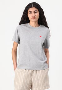 Gray T-shirt made of soft fabric, featuring a small red heart detail on the left chest. The model wears light-colored striped shorts.