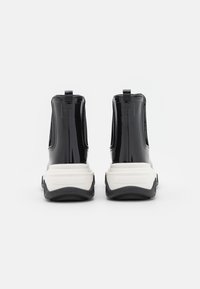 Par de botas de tobillo negras brillantes con suelas gruesas blancas, con lengüetas visibles y "Lemon Jelly" grabado en la parte trasera de los talones, vistas desde la parte trasera.