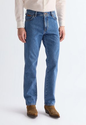 Man wearing light blue straight-leg jeans, beige shirt, and brown suede boots, standing against a white background.