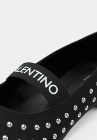 Sapatilha preta com parte superior de borracha, detalhes em prata com tachas e uma tira preta com o texto branco "VALENTINO". Textura suave em geral.