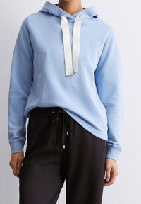 Felpa leggera blu con tasca frontale a marsupio, lacci bianchi e polsini a costine, abbinata a pantaloni jogger neri con cerniere laterali.