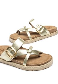 Metallisch goldene Sandalen mit Kreuzriemen und einer Schnallen-Detail. Weiche braune Innensohle, strukturierte Außensohle und gewebter juteähnlicher Besatz.