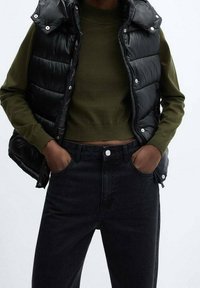 Czarna kamizelka puchowa o błyszczącej fakturze noszona na oliwkowo-zielonym obciętym swetrze, w połączeniu z czarnymi jeansami z wysokim stanem. Zapięcie na zatrzaski.