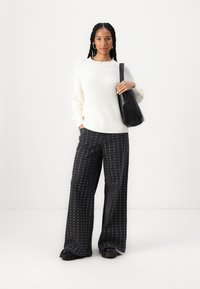 Weißer Strickpullover mit gerippter Textur, kombiniert mit schwarzen weit geschnittenen Hosen mit weißen Punkten. Das Model trägt eine schwarze Tasche.