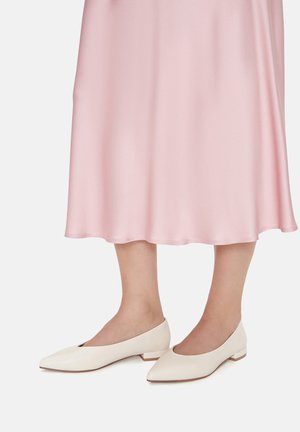 Untere Beine, die einen hellrosa Satin-Midirock und cremeweiße, niedriggeschossene spitze Schuhe tragen, stehend vor einem schlichten weißen Hintergrund.