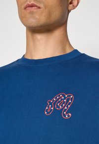 Camiseta azul de algodão com decote redondo, apresentando um logotipo bordado em vermelho com estrelas brancas, exibindo um tecido ribbed texturizado.