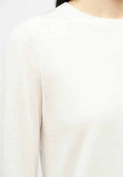 Sweater bege claro com uma textura suave, gola redonda e ombros sem costuras, exibindo um padrão de malha lisa.