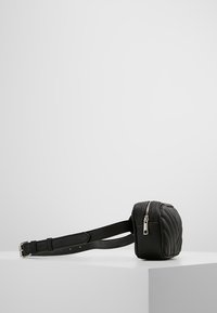 Musta tikattu nahkainen vyölaukku säädettävällä hihnalla ja hopeisella vetoketjulla, lepää valkoisella pinnalla vaaleanharmaata taustaa vasten.