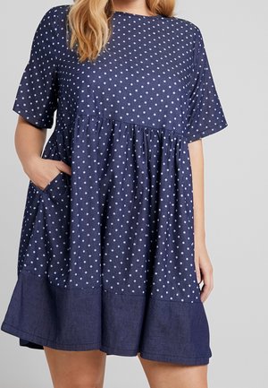 Kobieta ubrana w granatową sukienkę z krótkim rękawem w białe kropki, z jednolitym granatowym obszyciem na dole, z jedną ręką w bocznej kieszeni.