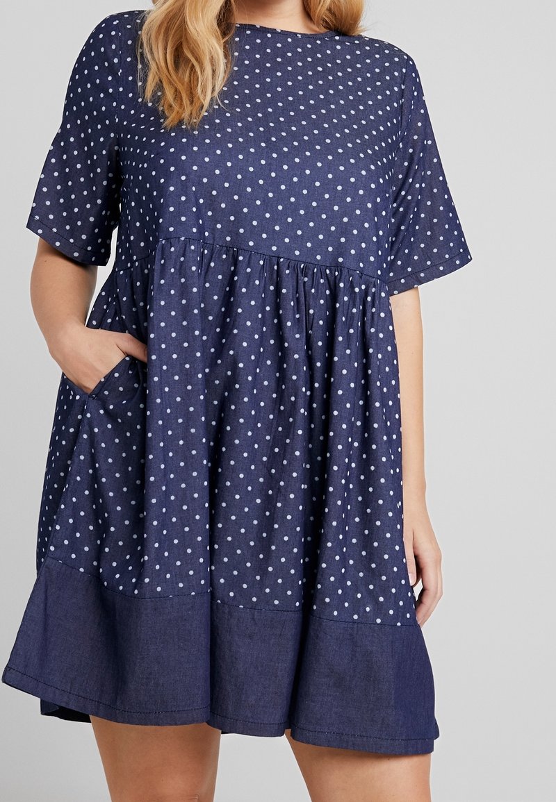 Femme portant une robe bleu marine à manches courtes avec des pois blancs et un ourlet uni bleu marine, une main dans la poche latérale.