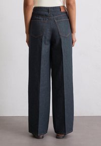 Leveälahkeiset farkut tummasävyisessä pesussa, joissa on sileä pinta, kaksi takataskua ja saumojen kontrastivärisenä oranssit ompeleet.