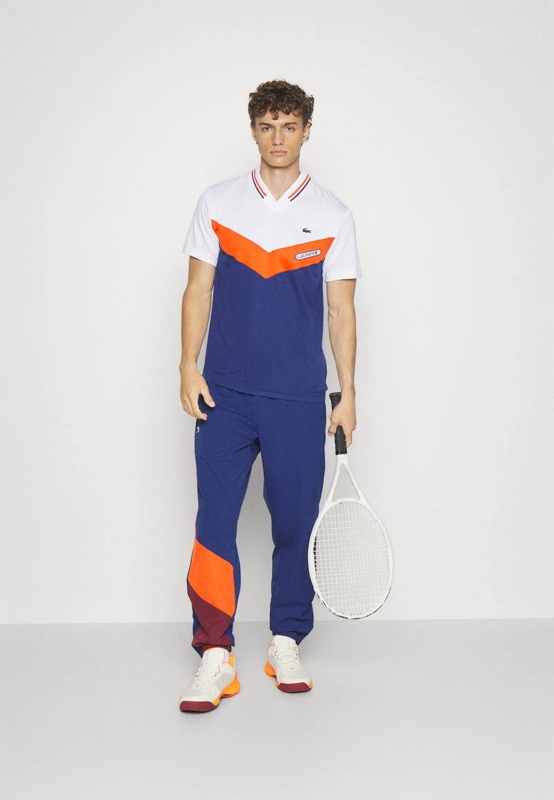 Uniforme de sport Lacoste bleu et blanc avec un motif en chevrons orange, associé à un pantalon marine avec des accents orange et bordeaux, tenant une raquette de tennis.