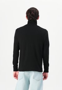 T-shirt noir à manches longues en tissu doux, avec col haut et silhouette ajustée. Vue de dos avec des coutures minimales, associé à un jean bleu clair.