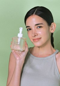 Przezroczysta kwadratowa butelka z olejkiem do demakijażu z pompką, oznaczona "NEEDLY", na zielonym tle. Butelka jest półprzezroczysta o miękkich krawędziach.