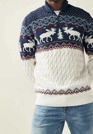 Mężczyzna ubrany w białą, grubą dzianinową sweter z niebieskimi i czerwonymi wzorami oraz białymi motywami łosi i drzew, w połączeniu z jasnoniebieskimi jeansami.