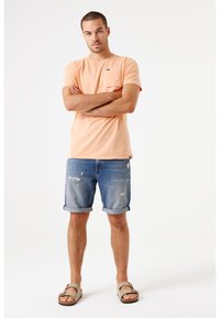 Camiseta corta de manga corta en color naranja claro con un bolsillo, combinada con pantalones cortos de mezclilla azul desgastados doblados en el dobladillo. Sandalias marrones completan el conjunto.