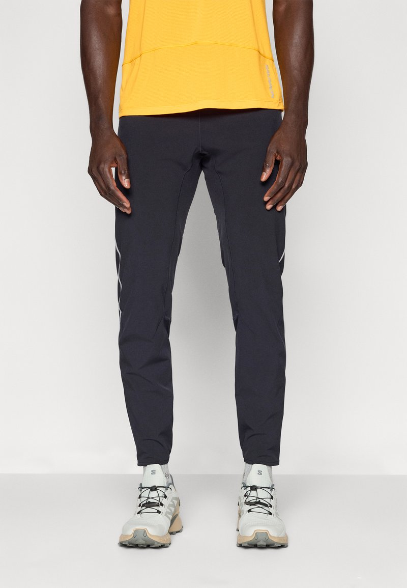 Homme portant un pantalon de sport noir, des chaussures de course blanches et beiges, et un t-shirt jaune à manches courtes sur un fond uni.