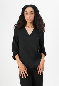 Zwarte V-hals blouse met lange puffmouwen, een gladde textuur en een relaxte pasvorm. G gedragen met zwarte broek, tegen een effen witte achtergrond.