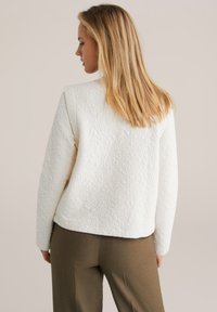 White textured sweater with raised patterned design, featuring a round neckline and a relaxed fit, paired with brown tailored trousers.