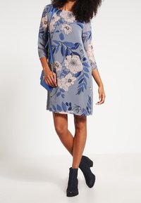 Floral-printed dress in gray, featuring three-quarter sleeves. The design includes large pink flowers and green leaves, complemented by ankle boots.