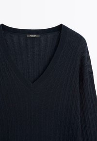 Maglione lavorato a maglia blu navy con scollo a V, texture a costine e motivo a treccia. Etichetta visibile nell'area del collo.