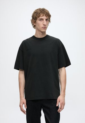 Junger Mann mit welligem braunem Haar, der ein lockeres schwarzes T-Shirt und schwarze Hose trägt, steht vor einem einfachen weißen Hintergrund.