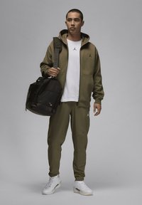 Oliven grøn hoodie og joggers med sorte accenter, hvid t-shirt med logo, bærende på en sort duffeltaske, stående mod en grå baggrund.