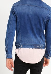 Chaqueta de denim en un color azul desvanecido, corte recto, cierre de botones y etiqueta de marca en la parte posterior. Superpuesta sobre una camiseta de color rosa claro.