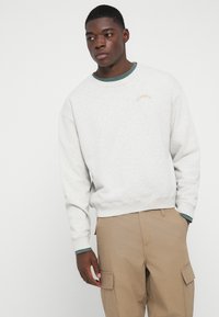 Sweatshirt gri deschis dintr-un material cu gât rotund, manșete ribă și un logo mic "Pompeii" în maro deasupra pieptului stâng.
