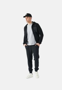Sudadera negra con cremallera, camiseta gris, pantalones jogger negros y zapatillas blancas. Una gorra negra complementa el atuendo. Textura de tela suave.