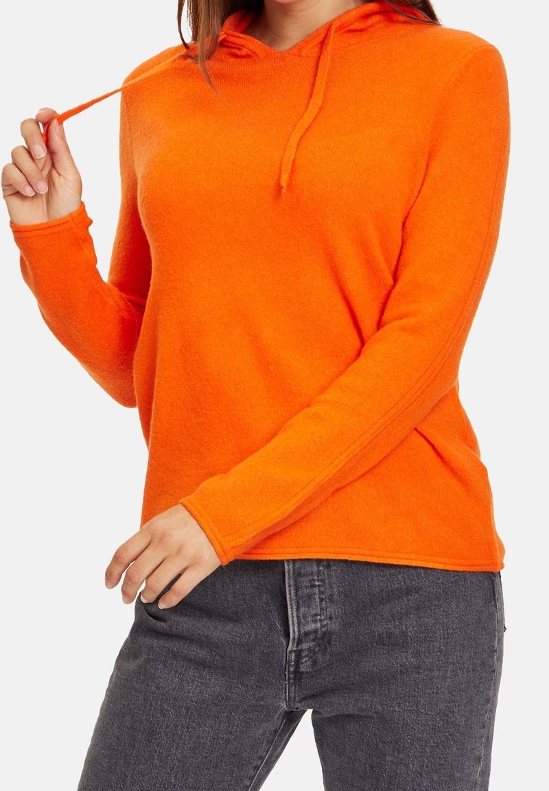 Femme portant un sweat à capuche orange vif et un jean gris foncé, tenant un cordon de la capuche avec sa main.