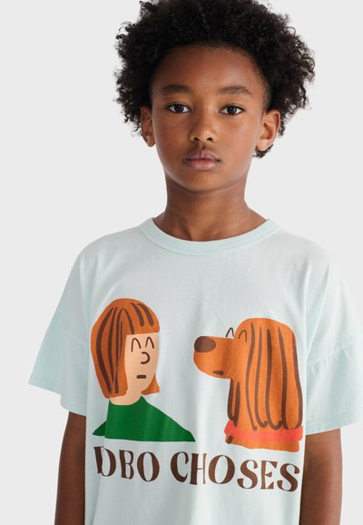 Dziecko z kręconymi włosami noszące białą koszulkę z nadrukiem kreskówkowej dziewczynki i psa zwróconych twarzami do siebie oraz napisem "DODO CHOSES" poniżej.