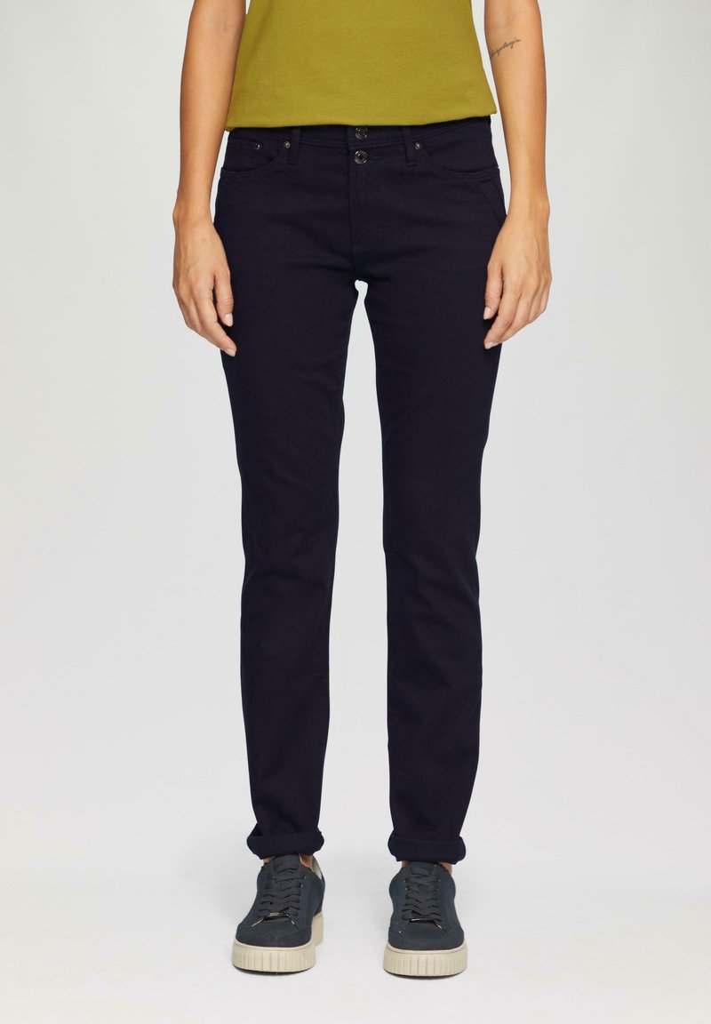 Temno mornarsko modre slim-fit kavbojke iz denima, ki vključujejo pet žepov, dvojno gumbno zapiranje in subtilne vezenine. V kombinaciji s sivimi športnimi copati.