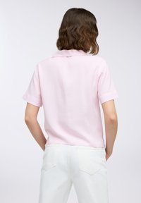 Camicetta rosa chiaro a maniche corte con colletto, caratterizzata da strisce verticali bianche. Indossata con pantaloni bianchi, che mostrano un fit rilassato.