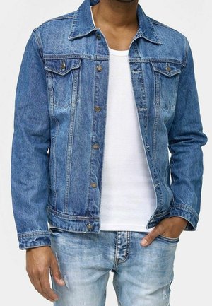 Mann trägt eine hellblaue Jeansjacke über einem weißen T-Shirt und dazu passende hellblaue Jeans, eine Hand in der Tasche, vor einfachem Hintergrund.