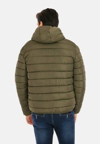 Uomo che indossa una giacca imbottita verde oliva con cappuccio, in piedi di spalle, abbinata a jeans blu scuro su uno sfondo bianco.