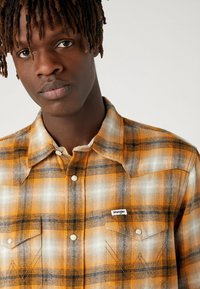Camicia in flanella a scacchi con colori arancione, nero e bianco. Presenta due tasche sul petto, bottoni a pressione in perla e un'etichetta Wrangler.