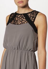 Graues Kleid aus fließendem Stoff, mit einem schwarzen, durchsichtigen Spitzenhalsausschnitt und schwarzen Akzentdetails an den Schultern. Mit elastischem Bund.