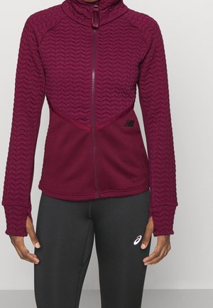 Vrouw draagt een bordeauxrode jas met volledige rits en gestructureerde mouwen en zwarte leggings met een wit logo op de dij, staand tegen een effen achtergrond.