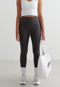Vrouw in een wit tanktop, hoge donkergrijze legging, witte sneakers en sokken, met een witte tote bag met zwarte tekst.