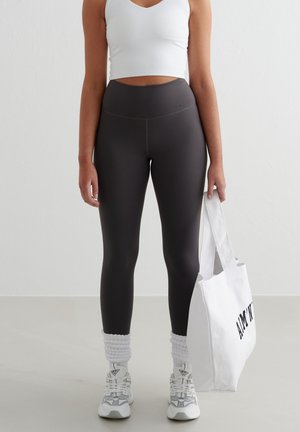 Vrouw in een wit tanktop, hoge donkergrijze legging, witte sneakers en sokken, met een witte tote bag met zwarte tekst.