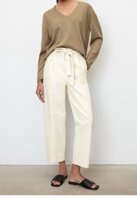 Maglione lavorato a maglia beige con scollo a V, abbinato a pantaloni beige a vita alta con laccio frontale. Sandali neri senza talone completano il look.