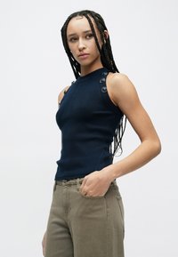 Top de alças azul-marinho com nervuras, botões nos ombros, modelo ajustado e tecido texturizado. Combinado com calças bege claro.