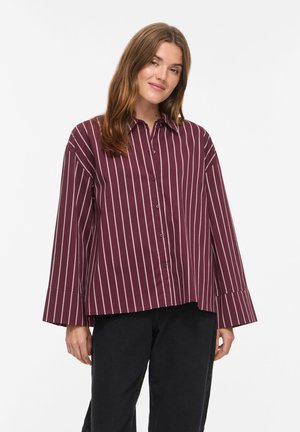 Femme aux longs cheveux bruns portant une chemise bordeaux ample à rayures verticales blanches et un pantalon noir, debout devant un fond blanc.