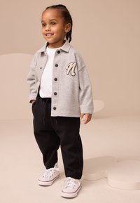 Chaqueta gris con botones y letra 'N' bordada, combinada con pantalones deportivos negros y zapatillas blancas. Diseño sencillo con colores contrastantes.