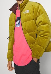 Grön kordfløjlsdunjacka med quiltad design, dragkedjestängning och hög krage. Bärs över en ljusrosa t-shirt med ett grafiskt tryck.