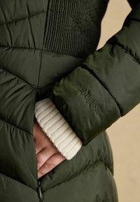 Zelená prešívaná bunda s textúrou, s bočným zipsom a krémovým rebrovaným manžetovým nápletom. Na rukáve je embosované logo značky "JOULES".