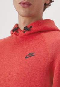 Närbild av en person som bär en röd Nike-huvtröja med svarta dragkedjeproppar och en svart Nike-logga på bröstet.