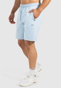 Pantalones cortos deportivos de color azul claro con cinturilla ajustable, bolsillos laterales, textura suave y una pequeña etiqueta de marca en la parte frontal. Combinados con zapatillas blancas.