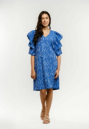 Kobieta ubrana w niebieską sukienkę za kolano z fakturowanym wzorem liści oraz krótkimi falbankowymi rękawami, w beżowych sandałach na paski, stojąca.